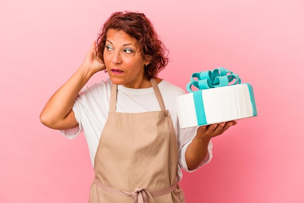 Mulher latina pastelaria de meia idade segurando um bolo isolado no fundo rosa, tentando ouvir uma fofoca.