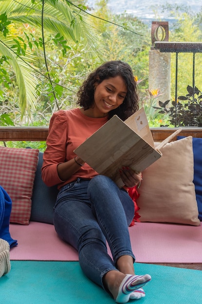 Mulher latina sorrindo casualmente vestida na sala de estar, apoiando-se em almofadas coloridas enquanto lê um livro