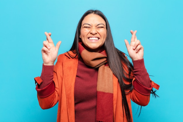 Mulher latina sorrindo e cruzando os dedos ansiosamente, preocupada e desejando ou esperando por boa sorte