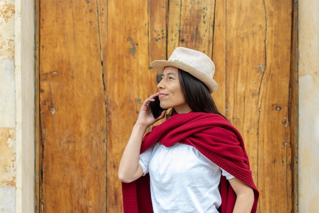 Mulher latina usando chapéu e um xale vermelho enquanto fala ao telefone com uma velha porta de madeira ao fundo