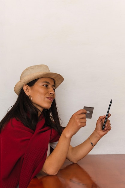Mulher latina usando chapéu e um xale vermelho sentada segurando seu telefone celular e um cartão de crédito em fundo branco