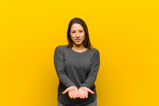 Mulher latino-americana, sorrindo alegremente com olhar amigável, confiante e positivo, oferecendo e mostrando um objeto ou conceito isolado contra a parede amarela