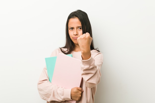 Mulher latino-americano nova que guarda alguns cadernos que mostram o punho à câmera, expressão facial agressiva.