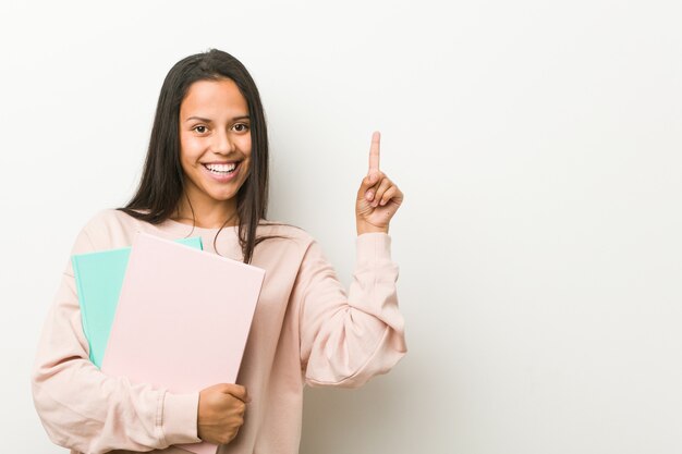 Mulher latino-americano nova que guarda alguns cadernos que sorriem alegremente apontando com o dedo indicador afastado.