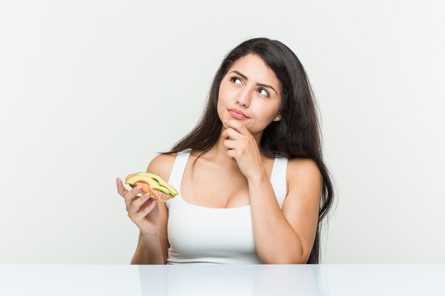 Mulher latino-americano nova que guarda um brinde do abacate que olha lateralmente com expressão duvidosa e cética.