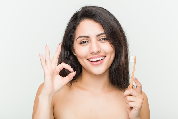 Mulher latino-americano nova que mantém uma escova de dentes alegre e segura mostrando o gesto aprovado.