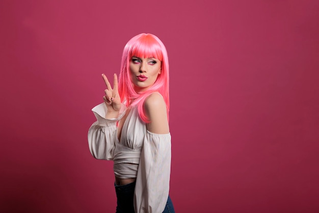 Mulher legal na moda com cabelo rosa fazendo sinal de paz e beijo na frente da câmera. garota atraente despreocupada mostrando símbolo de vitória e gesto pacífico com dois dedos no estúdio.