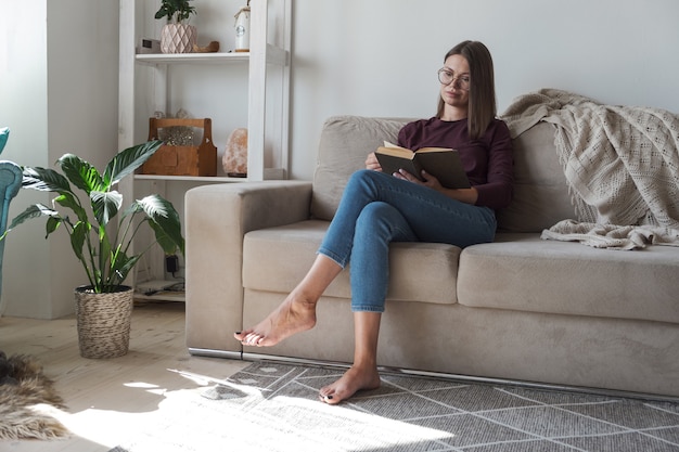 Mulher lendo livro e relaxando sentada no sofá em casa