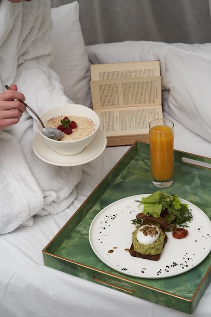 Mulher lendo livro enquanto toma café da manhã na cama. mulher de roupão tomando café da manhã na cama do hotel, close-up