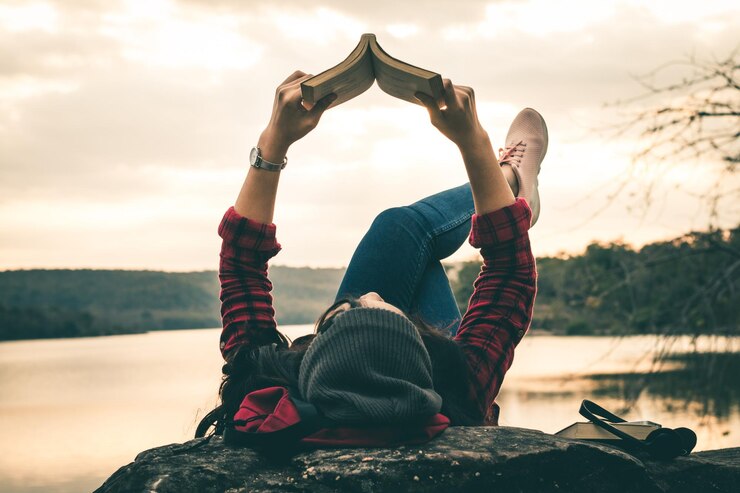 Foto mulher lendo um livro deitada na beira do lago durante o pôr-do-sol