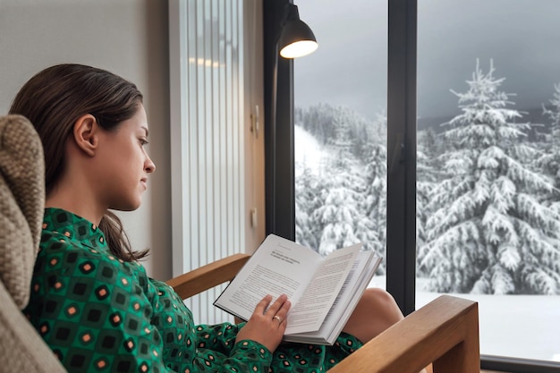Mulher lendo um livro em casa aconchegante perto da janela com vista panorâmica de inverno