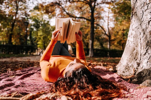 Foto mulher lendo um livro enquanto está deitada no parque durante o outono