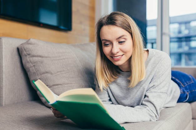 mulher lendo um livro interessante deitada no sofá, descansando depois do trabalho