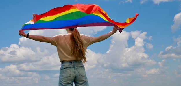 Mulher lésbica bissexual transgênero segura bandeira LGBT contra o céu azul com nuvens em um dia ensolarado e celebra o dia da bissexualidade ou o dia nacional de sair do armário