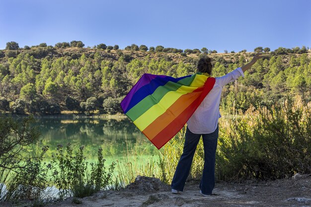 Mulher lésbica de meia-idade irreconhecível segurando a bandeira gay do arco-íris no lago ao ar livre. Conceito de liberdade