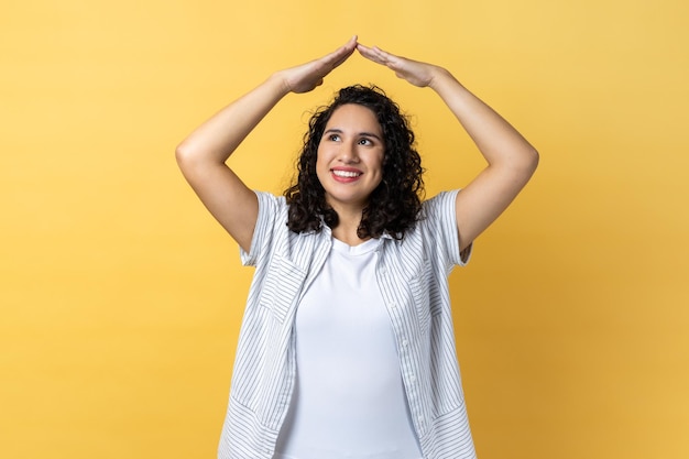 Mulher levantando as mãos mostrando o gesto do telhado e sorrindo contente sonhando com a casa olhando para longe