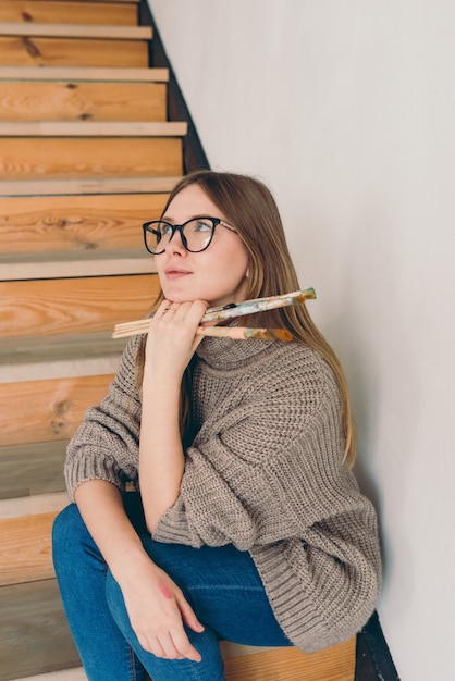 Mulher linda artista de óculos e roupa casual sentada na escada com escovas