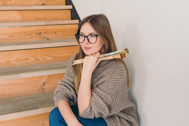 Mulher linda artista de óculos e roupa casual sentada na escada com escovas