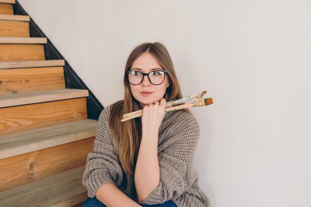 Mulher linda artista de óculos e roupa casual sentada na escada com escovas