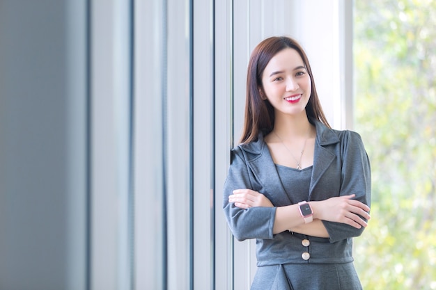Mulher linda asiática com cabelo comprido usa um vestido cinza e os braços cruzados enquanto fica perto da janela.
