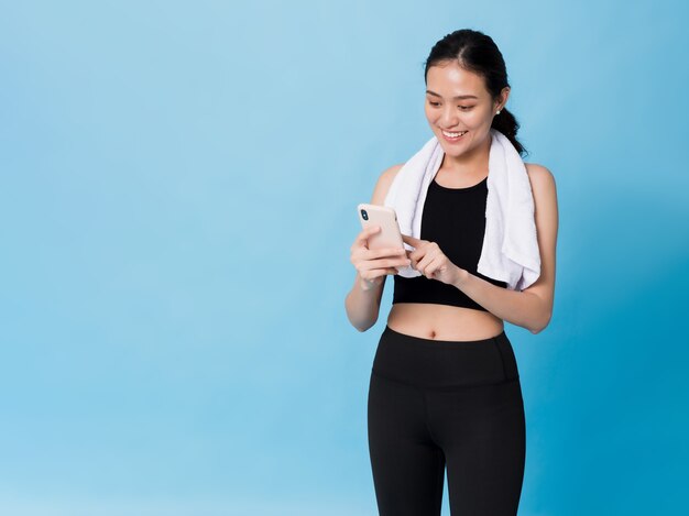 Mulher linda asiática feliz usando smartphone após exercício isolado em azul