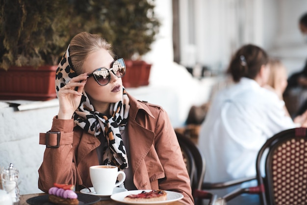 Mulher linda bebendo café em um café vestindo uma jaqueta elegante, óculos de sol e lenço de seda ao ar livre