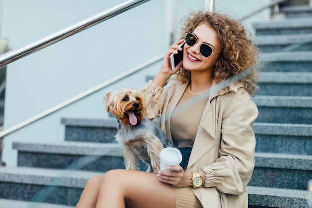 Mulher linda com cabelos cacheados em roupas da moda sentar na cidade com seu cachorro e falando ao telefone.