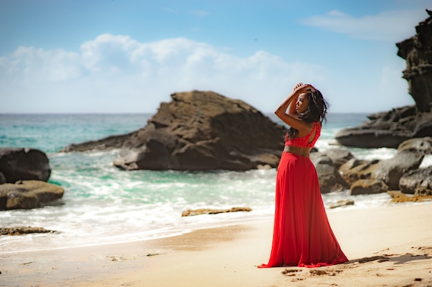 Mulher linda e sensual usando um vestido vermelho luxuoso e posando ao lado de um penhasco de areia