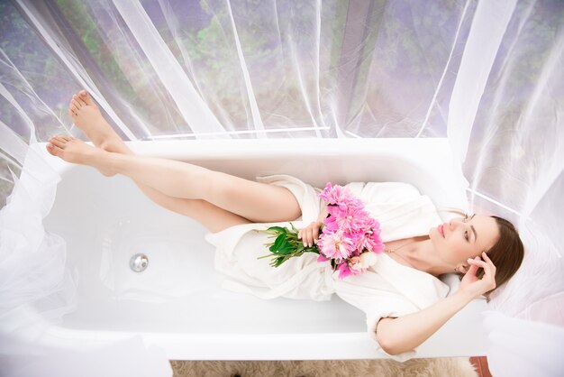 Mulher linda em um roupão branco com um buquê de peônias rosa perto da banheira