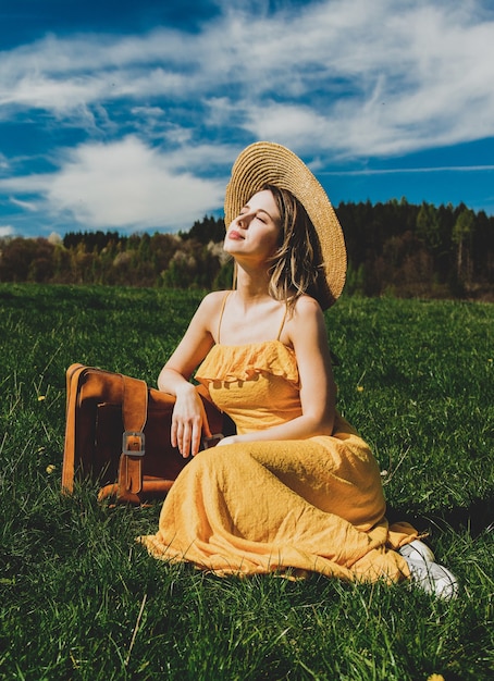 Mulher linda em um vestido amarelo e uma mala sentada num prado de montanha com flores