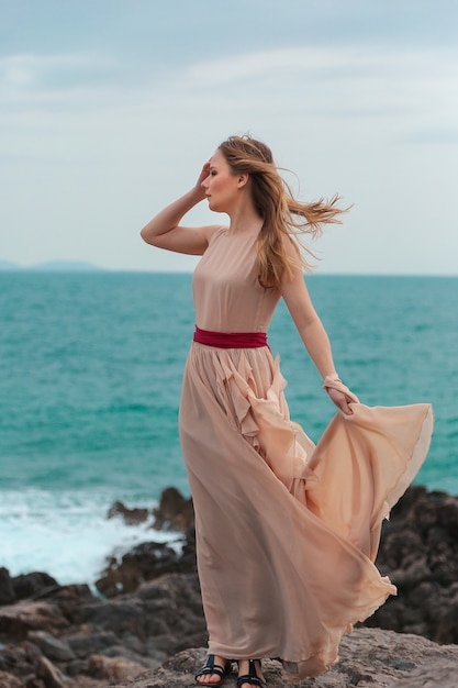 Foto mulher linda em um vestido bege fica em uma praia rochosa