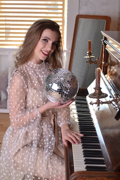 Mulher linda em um vestido bege segura uma bola de discoteca na mão e toca um piano retrô