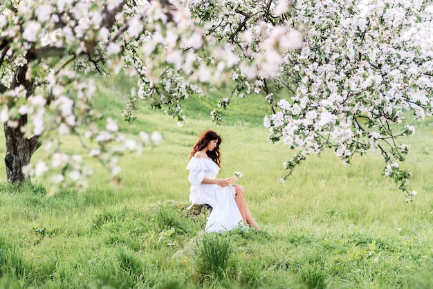 Mulher linda em um vestido branco lê um livro no jardim primavera. Uma garota no fundo de árvores floridas.