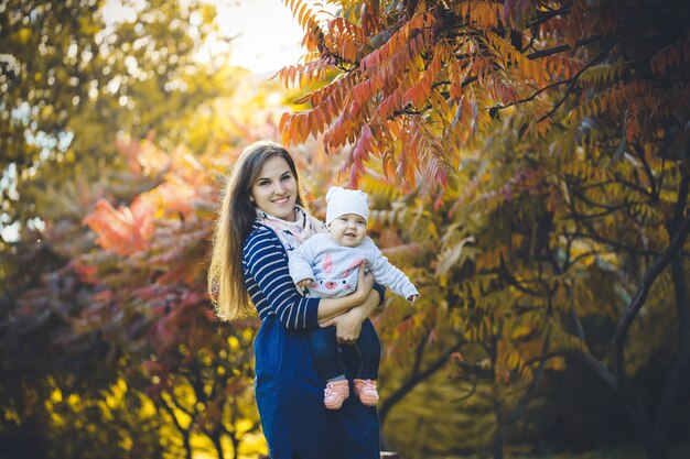 Mulher linda em um vestido brinca com uma criança nos braços, de pé na natureza outono com folhas caídas. Jovem garota de cabelos compridos relaxar no parque com folhas vermelhas amarelas no outono. Família feliz.