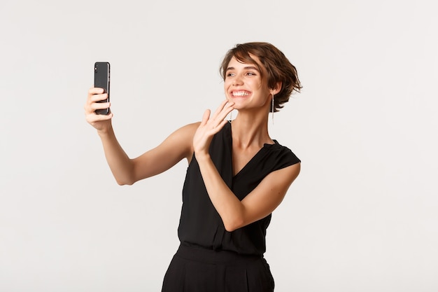 Mulher linda feliz tendo vídeo chamada, acenando com a mão para a câmera do smartphone e sorrindo, branco de pé.