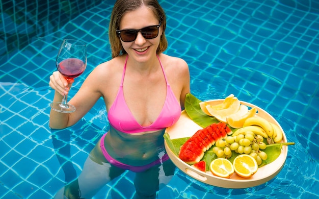 Mulher linda na piscina com um copo de vinho e uma bandeja flutuante de frutas