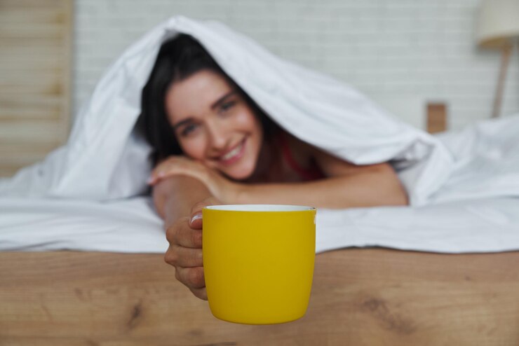 Foto mulher linda olhando para fora de um cobertor e estendendo a mão com a xícara enquanto estava deitada na cama