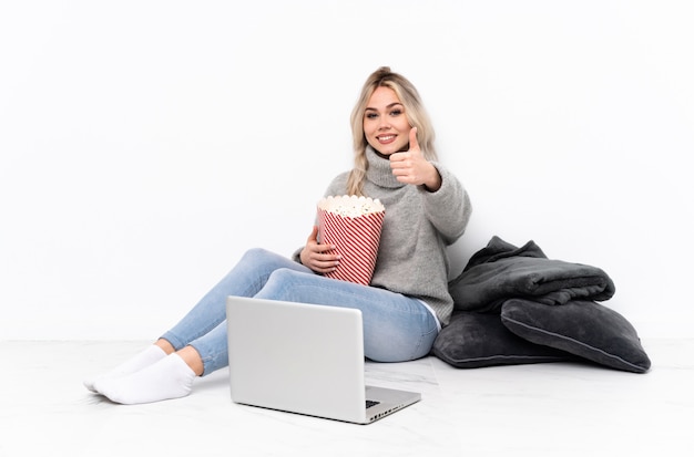 Mulher loira adolescente comendo pipoca enquanto assiste a um filme no laptop com polegares para cima, porque algo de bom aconteceu