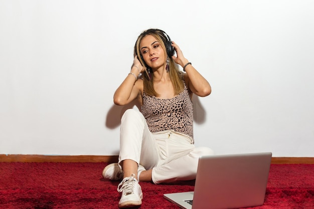 Mulher loira adulta usando seus fones de ouvido e seu laptop ouvindo música sentado em um tapete vermelho e contra um fundo branco