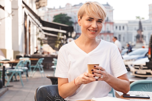 mulher loira alegre de camiseta branca sentada em um café de rua no verão e bebendo café em um copo de papel