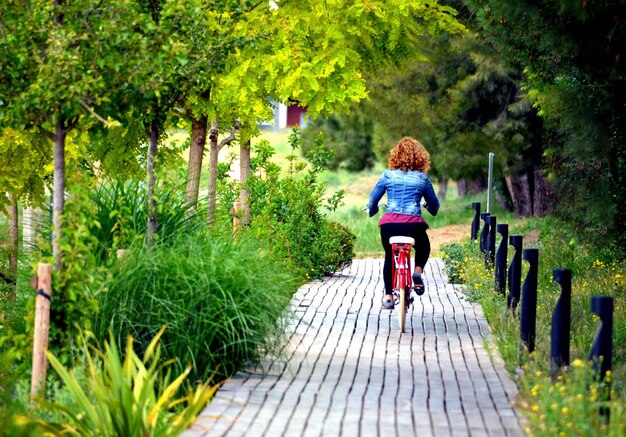 Mulher loira andando no parque com um jeans azul em uma bicicleta vermelha