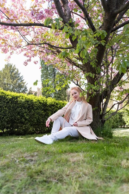 Mulher loira atraente sentada em um parque em flor no gramado caloroso dia de primavera garota atraente em repouso quadro vertical