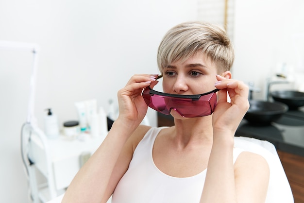 Mulher loira atraente sentada na cadeira médica na sala de cosmetologia do salão spa e colocando óculos de proteção UV
