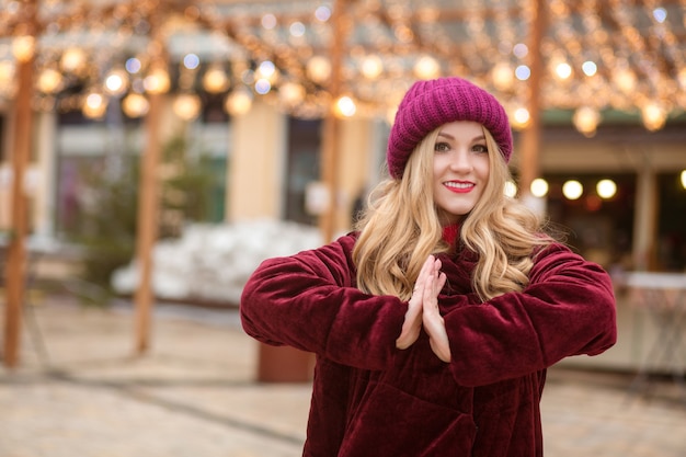 Mulher loira atraente usando chapéu de malha vermelho e casaco de inverno, posando no fundo de uma guirlanda na rua em Kiev