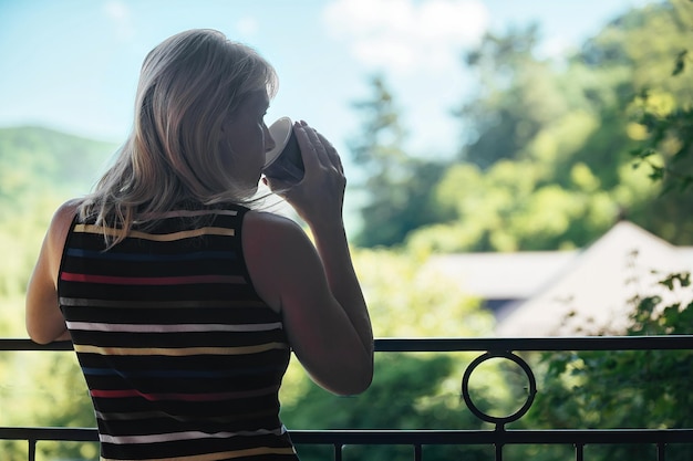 mulher loira bebe café no início da manhã na varanda contra o pano de fundo da natureza
