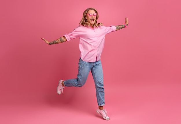 Mulher loira bonita com camisa rosa e jeans sorrindo pulando na rosa isolada se divertindo