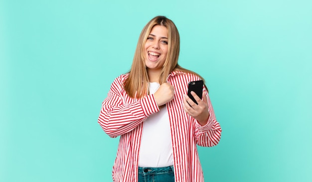 mulher loira bonita curvilínea se sentindo feliz e enfrentando um desafio ou comemorando e segurando um telefone inteligente