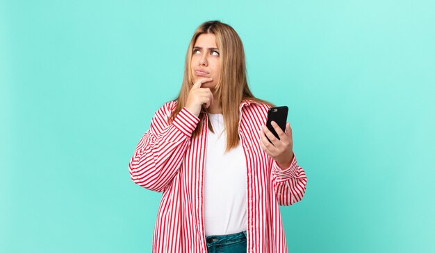 Mulher loira bonita e curvilínea pensando, se sentindo em dúvida e confusa e segurando um telefone inteligente