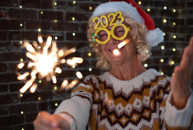 Mulher loira bonita sênior com óculos engraçados e suéter de inverno segurando a luz do faísca idosa feliz celebrando o Natal e o ano novo brilhando na noite