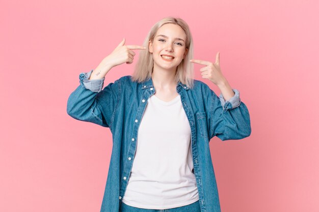 mulher loira bonita sorrindo com confiança apontando para o próprio sorriso largo